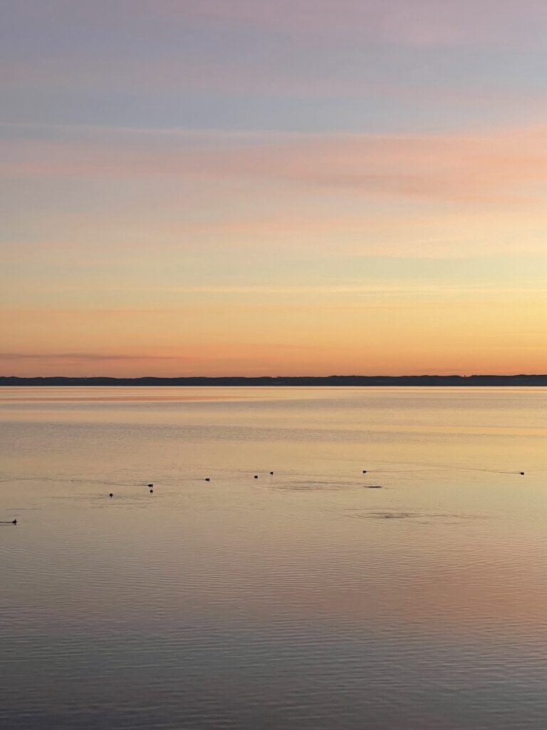 Farbintensive Abendstimmung über spiegelndem See und Horizont in der Ferne.