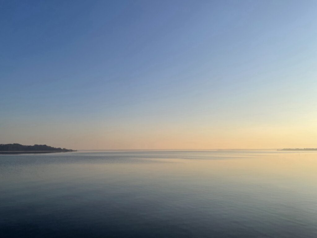 Weite, ruhige Wasserfläche unter blauem Himmel mit warmem Licht am fernen Horizont.